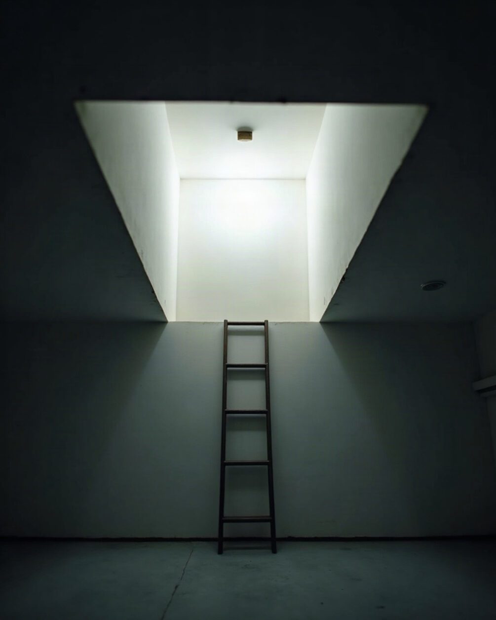 Loft hatch open in a ceiling with a ladder edge visible and dim light beyond, plain landing below