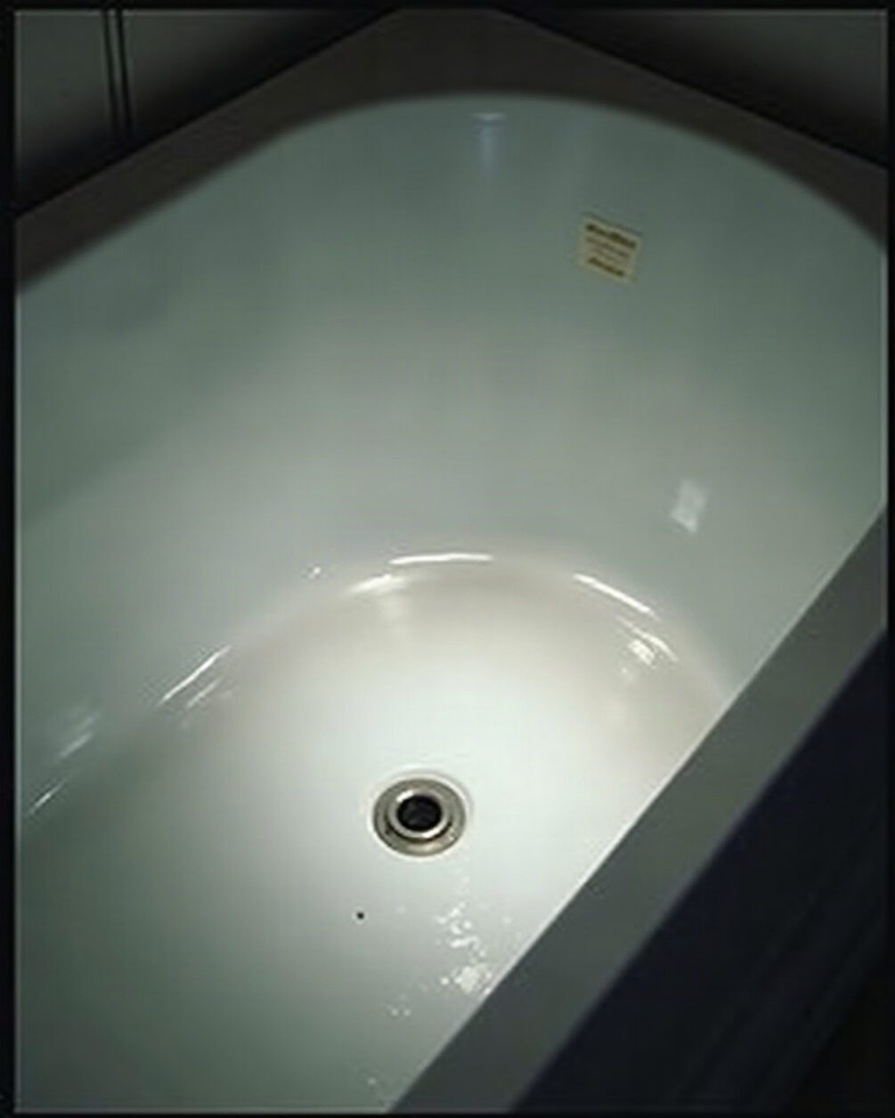 Empty white bath seen from above under an overhead light, smooth enamel sides and the drain in shadow
