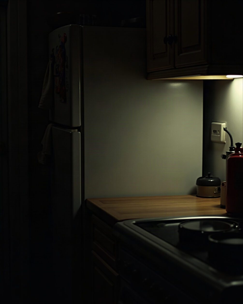 Kitchen at night with a light just switched on, worktop edge and the shadowed gap behind a fridge